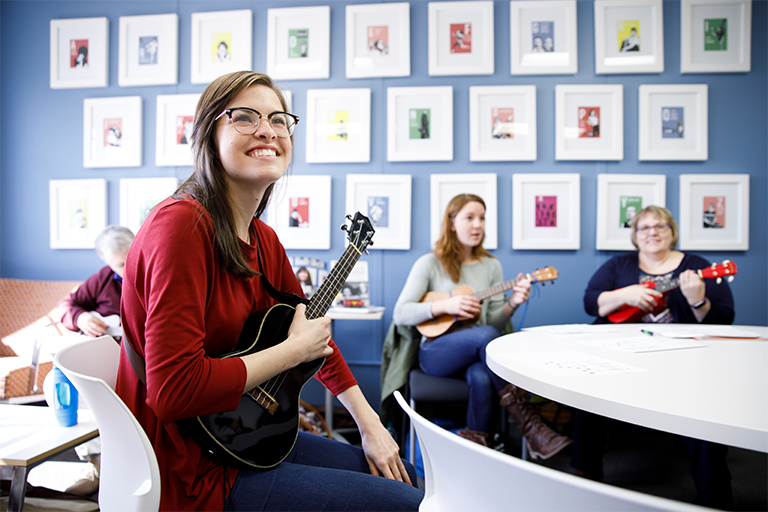female student with a red shirt playing a ukulele. There are two more females holding ukuleles in the background. 