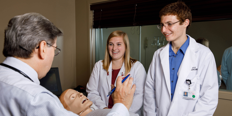 Two medical students smile as their professor explains something over a teaching mannequin.